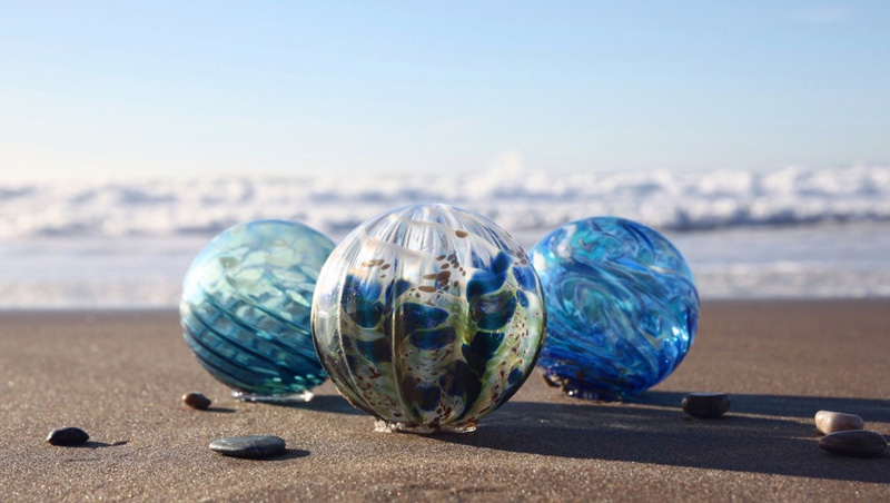 Three beautiful glass floats in blue colors, on the sand in front of the ocean surf.