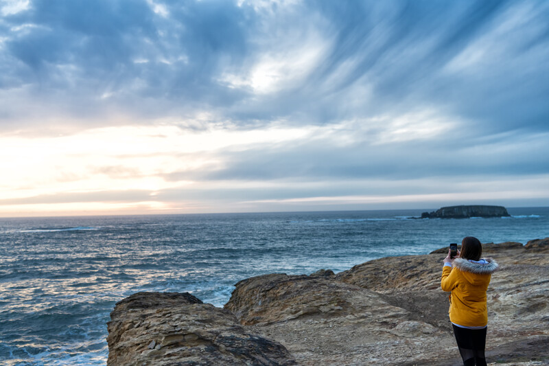 Girl taking photo at sunset with blue cloudy sky on an Oregon Coast rocky shore.