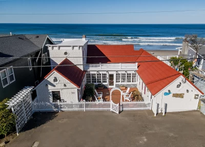 Aerial view of beautiful a white classic beach house with a reddish roof, white picket fenced surrounding a courtyard, arched trellis entrance, and oceanfront views.