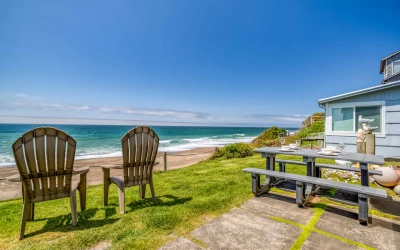Picnic table with oceanfront views, 5 fancy place settings on a green grass yard, and 2 Adirondack chairs..
