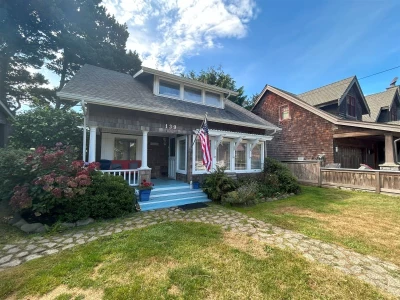 Exterior view of a beautiful 2-level cedar shake cottage busting with cottage style charm. A manicured grass yard with a stone walkway leading to the front porch.
