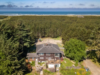 Aerial view of a 3-level large house surrounded by fir trees with the Pacific ocean in the distance.