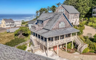 Aerial view of a large 3-level gray cedar shake beach house with a wrap-around covered deck, beautiful cream colored craftsmen style railings, and a distant ocean view.