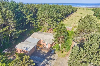 Aerial view of single level salmon colored house, with a large area of treed land is cleared leading to the ocean.
