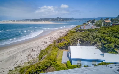 Aerial view of a flat roof beach house on a small cliff, blue ocean, sandy beach, and views of the Alsea Bay Bridge.