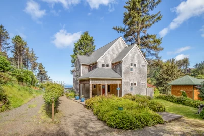 Exterior of tall 3 story beach house with gray shingles, a large covered porch, high ceiling roof lines, and landscaping.