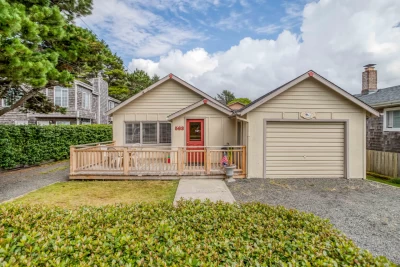 Tan colored beach house with an orange front door, single car garage, fenced front deck, and walkway to their small cottage..