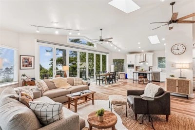 Huge, white, open kitchen-living room space with a wall of floor to ceiling windows. Vaulted ceiling, 2 fans, table and tan sofa.
