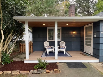 Covered porch with lighting, 2 chairs with pillows on a rug, and fresh landscaping.