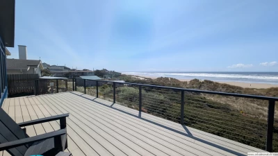A large beachfront deck with a black Adirondack chair overlooking low lying dunes, a sandy beach, and the ocean on a blue sky day.