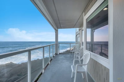 Four white outdoor chairs on a condo balcony with a glass railing, up-close oceanfront views with a rock barrier.
