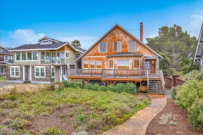 Two-level house with fresh cedar shake siding, front yard of shrubs, a sidewalk up a large deck, pointed roofline, and a lot of windows.