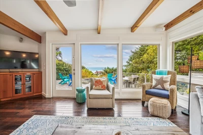 Living room view with big screen TV, floor to ceiling windows looking out onto the deck with an ocean view carved out around the shrubbery.