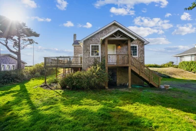 Cedar shake 2-level cottage style house with stairs leading up to the front door, a deck on the opposite side, surrounded by a grassy yard.